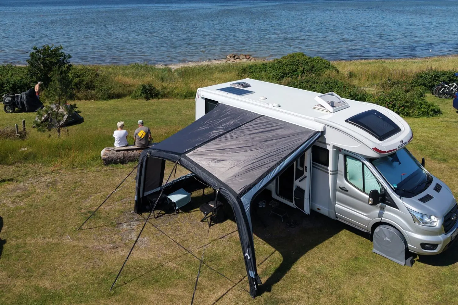 Motorhome with awning on a panorama pitch and two guests on a bench overlooking the Limfjord