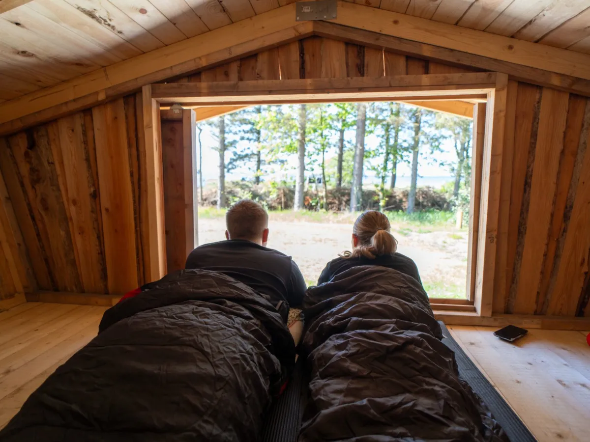 Stel in slaapzakken kijkt vanuit de shelter uit op het bos en de Limfjord